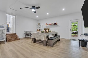 Un comedor moderno con una mesa preparada para cuatro, suelo de madera, ventilador de techo y vistas a la vegetación a través de una puerta.