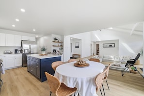 Une cuisine moderne avec des meubles blancs, un îlot central bleu et un coin repas doté de chaises en osier et d’une nappe blanche.