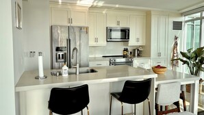 A modern kitchen with white cabinetry, stainless steel appliances, a central island with bar stools, and a large window providing natural light. - Sundrenched 2 BDRM waterfront gem with lake view near CNE, BMO Field (Toronto)