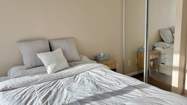 A bedroom with a bed, pillows, a bedside table, a mirror, and a wardrobe. - Sundrenched 2 BDRM waterfront gem with lake view near CNE, BMO Field (Toronto)