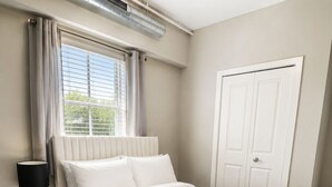 A bedroom with a bed, a nightstand, a lamp, a window with curtains, a radiator, and a door. - Cozy 1-bedroom apartment in serene Atlanta with fitness room (Atlanta)