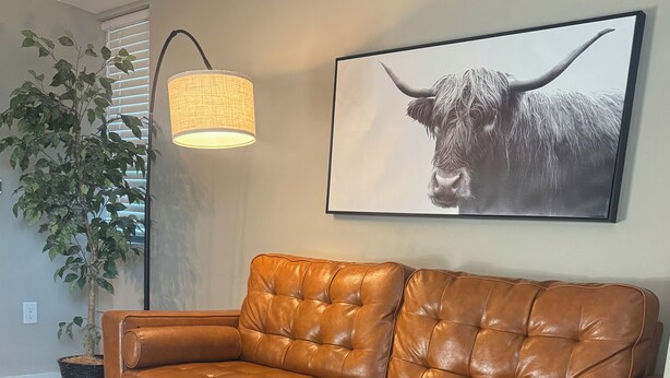 A brown leather sofa with a tufted backrest, a round coffee table with a plant and a book, a wall-mounted lamp, and a framed picture of a bull. - Cozy 1-bedroom apartment in serene Atlanta with fitness room (Atlanta)