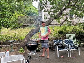 Una persona in piedi accanto a un barbecue all'aperto, con sedie e un edificio sullo sfondo.