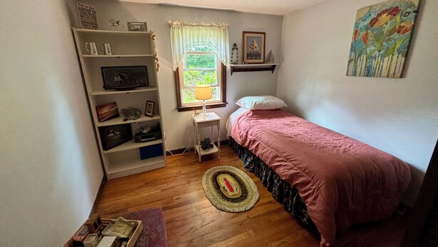 A small bedroom with a single bed, a nightstand, a shelf with various items, a window with curtains, and a painting on the wall. - Enjoy the best of Cape Cod. Perfect. Private.Parking. Pets. Available Sept 1 (Dennis)