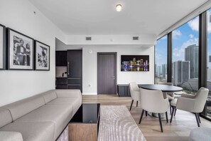 A modern living room with a sofa, a coffee table, and a dining area with chairs. There is a door leading to a kitchen and a large window with a city view. - Miami Pulse With Resort Amenities (Miami)