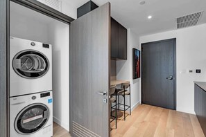 A modern laundry room with stacked washers and dryers, a bar stool, and a dark door. - Miami Pulse With Resort Amenities (Miami)