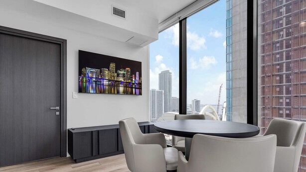 A modern room with a round table, four chairs, a cityscape painting, and a large window with a view of buildings. - Miami Pulse With Resort Amenities (Miami)