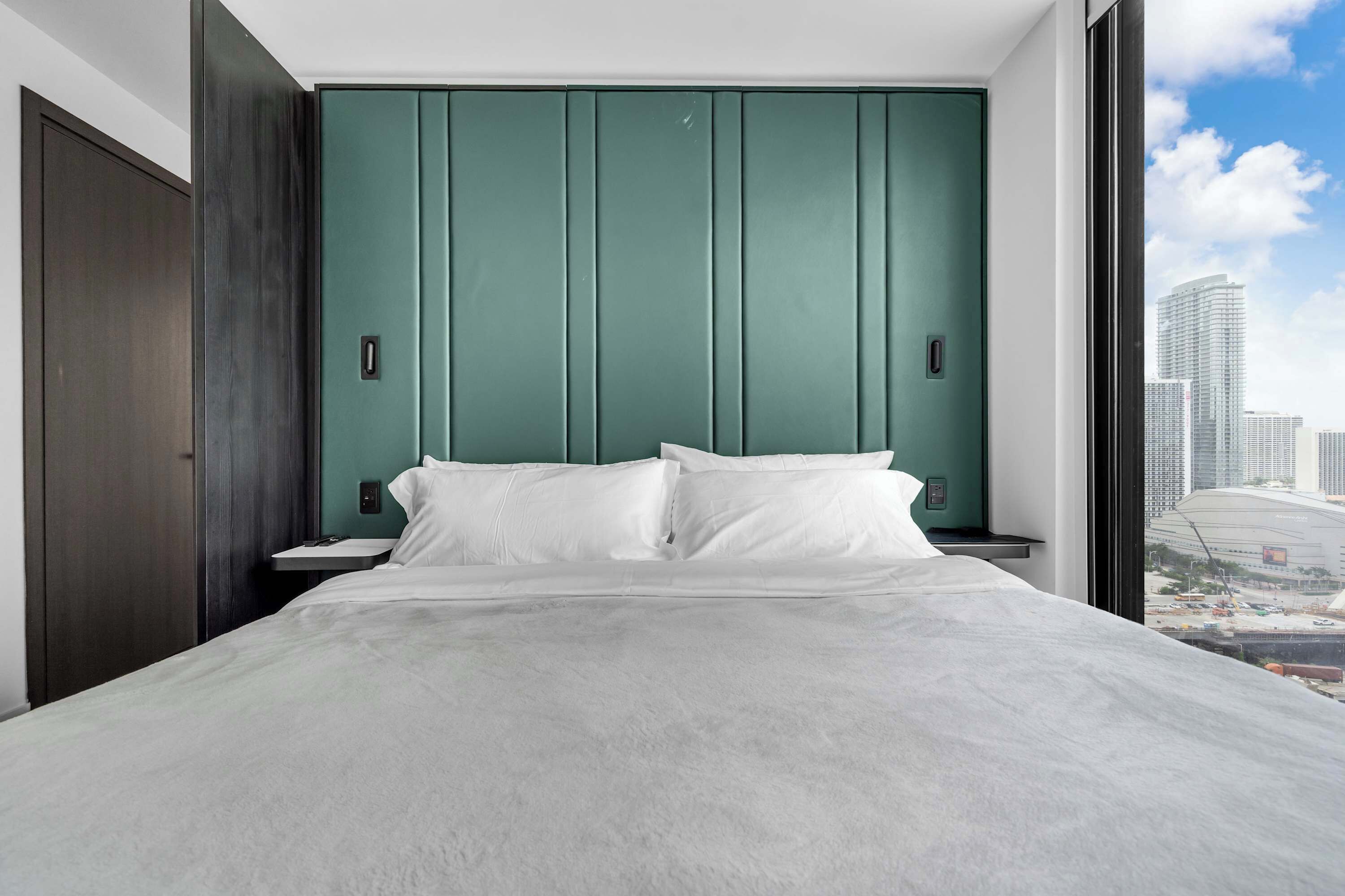 A modern hotel room with a large bed, a green headboard, and a view of the cityscape.