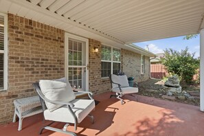 A covered patio with two white cushioned chairs and a wicker bench, a brick wall, and a wooden fence in the background. - Welcome to The Amsterdam Place. (Madison)