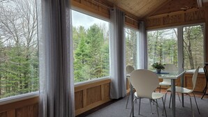 A room with large windows, wooden walls, a dining table with chairs, and a potted plant.