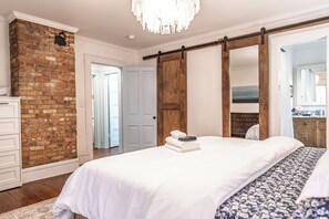 Une chambre avec un mur en briques, un lit avec une literie blanche, une porte en bois et un lustre.
