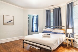 A bedroom with a metal bed frame, a bedside table with a lamp, and a window with curtains. - NEW! Downtown Charmer- Steps to downtown SoPines! (Southern Pines)