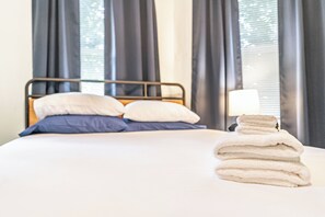 A neatly made bed with pillows, a stack of towels, and a bedside lamp in a room with large windows and curtains. - NEW! Downtown Charmer- Steps to downtown SoPines! (Southern Pines)