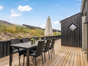 En træterrasse med et bord og stole, en hvid parasol og udsigt til strand og klitter.