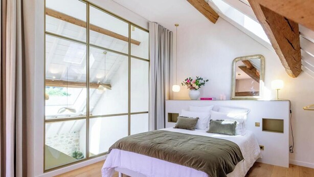 Quarto com uma janela grande, cama com roupa de cama branca, teto de madeira e mesa de cabeceira com abajur.