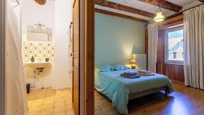 A bedroom with a bed, a bedside lamp, a window with curtains, and a bathroom with a sink and mirror. - The Priory Gardens (CLERY)