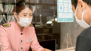 A hotel reception area with a staff member in a pink uniform behind a counter, a customer in a black jacket, and a menu board with various options. - Toyoko Inn Mito Eki Minami Guchi (Mito)