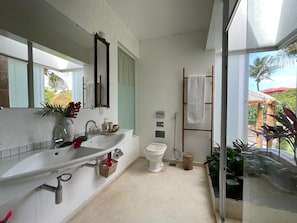 Un baño moderno con un espejo grande, un lavamanos blanco y un inodoro. Hay un perchero con una toalla colgada, y una ventana que ofrece vistas al exterior.