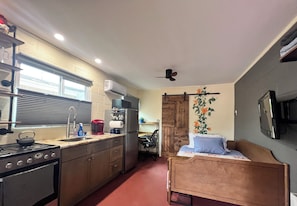 Eine kleine Küche mit Herd, Spüle und Kühlschrank, ein Essbereich mit einem Stuhl und ein Bett mit einem Holz-Kopfteil.