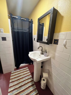 Ein Badezimmer mit weißem Waschbecken, einem Spiegel, einer Duschvorhang, einem Streifen Teppich und einem Toilettenpapierhalter.