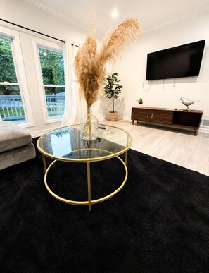 A modern living room with a glass coffee table, a flat-screen TV mounted on the wall, a wooden entertainment unit, and a view of the outdoors through large windows. - 2-bedroom house in peaceful East Point Atlanta (East Point)