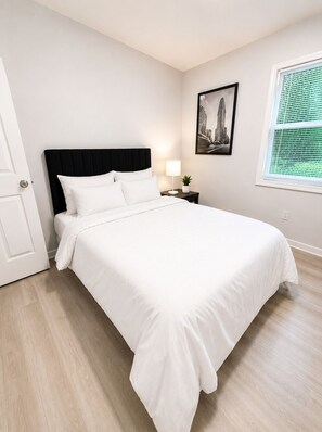 A neatly made bed with white bedding, a black headboard, and a small bedside table with a lamp and a plant. - 2-bedroom house in peaceful East Point Atlanta (East Point)