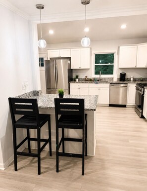 A modern kitchen with white cabinets, stainless steel appliances, a granite countertop, and two black bar stools. - 2-bedroom house in peaceful East Point Atlanta (East Point)