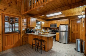 A wooden cabin kitchen with a central island, bar stools, a refrigerator, and a microwave. - Family Cabin ✨️ Hot Tub ✨️ Game Room ✨️ 2 Mi to The Island✨️ 5 Mi to Dollywood (Pigeon Forge)