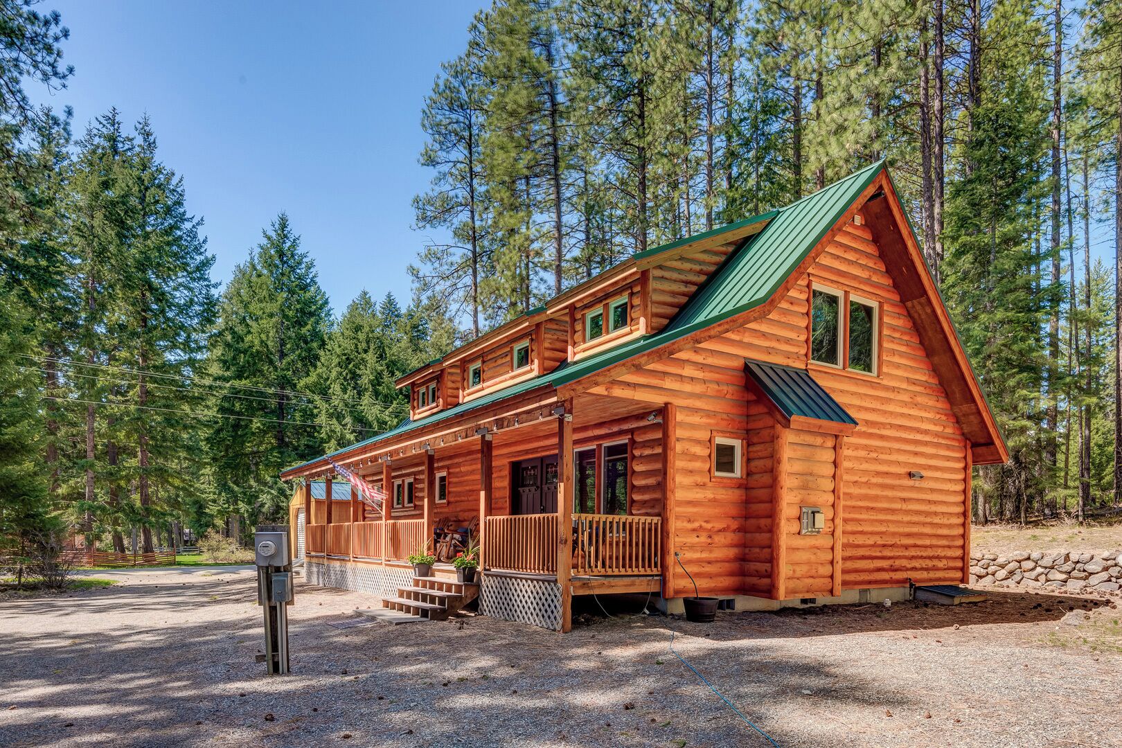 A log cabin with a porch, multiple windows, and a green roof, surrounded by tall trees.
