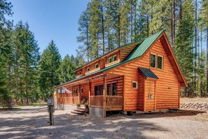A log cabin with a porch, multiple windows, and a green roof, surrounded by tall trees. - Beautiful home nestled in the forest walking distance to Lake Cle Elum/Beach. (Ronald)