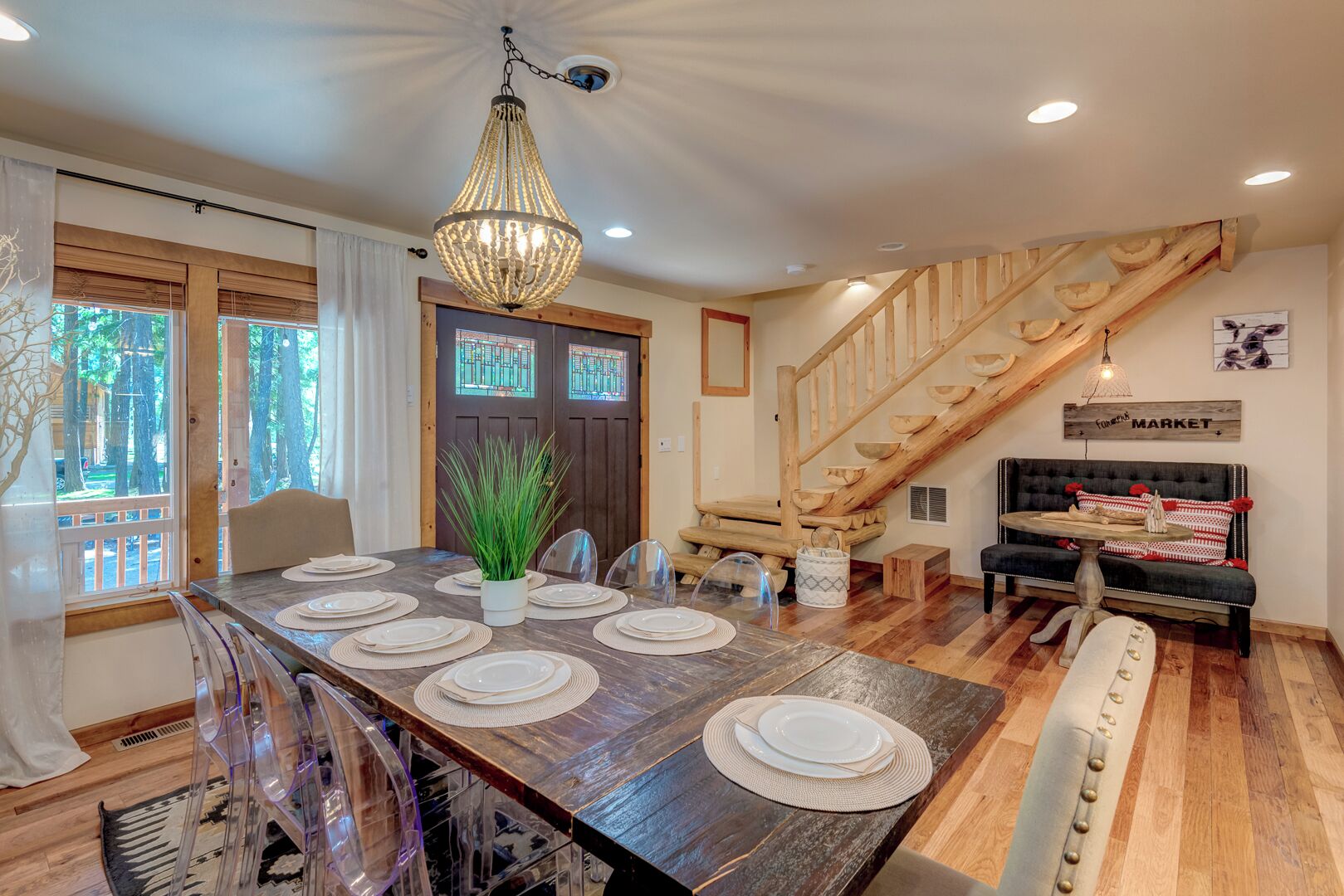 A dining area with a wooden table set for six, a chandelier, a staircase with wooden railings, and a sofa with patterned cushions.