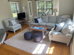 A living room with a sofa, coffee table, ottomans, and a TV. - Charming 4-bedroom house in Miacomet (Nantucket)