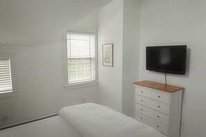 Une chambre à coucher comprenant un lit, une commode, un téléviseur fixé au mur et une fenêtre avec des stores.