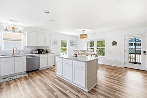 A modern kitchen with white cabinetry, a central island, and a dining area with a view. - Family & Dog Friendly Retreat w. Beach, Dog, and Baby Gear. Screened porch! (Oak Island)