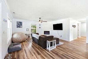 A modern living room with a sofa, ottoman, and bean bag chair, a flat-screen TV mounted on the wall, and a ceiling fan. - Family & Dog Friendly Retreat w. Beach, Dog, and Baby Gear. Screened porch! (Oak Island)