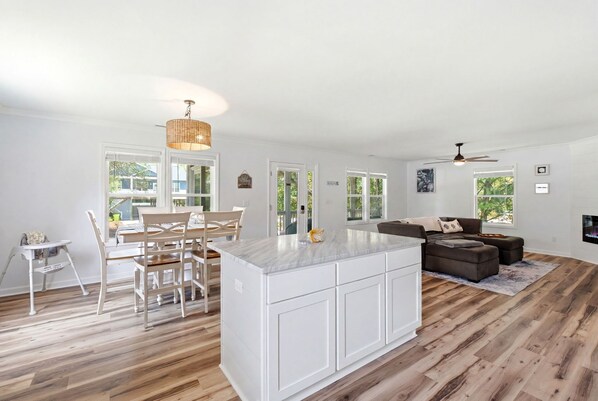 A spacious kitchen with a central island, white cabinetry, and a dining area with wooden chairs and a table. - Family & Dog Friendly Retreat w. Beach, Dog, and Baby Gear. Screened porch! (Oak Island)