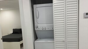 A compact laundry room with a washing machine and dryer, a small seating area with a bench, and a door leading to another room. - Modern unit with premium finish, porcelain floor and rain fall shower (Hollywood)