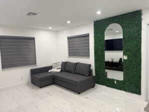 A modern living room with a grey sectional sofa, two windows with blinds, a green wall with a mirror, and a tiled floor. - Modern unit with premium finish, porcelain floor and rain fall shower (Hollywood)