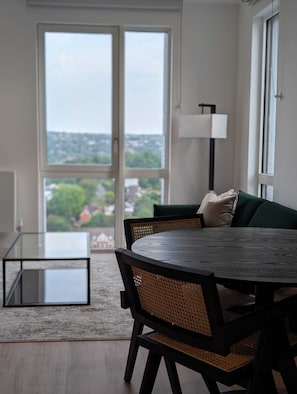 Ein modernes Wohnzimmer mit rundem Tisch, zwei Sesseln, einer Couch, einer Stehlampe und einem großen Fenster mit Blick auf die Stadt.