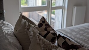 Un lit avec une literie blanche et des oreillers à motifs, une fenêtre avec des stores et une vue sur la verdure à l’extérieur.