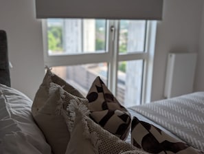 Ein Bett mit weißer Bettwäsche und gemusterten Kissen, ein Fenster mit Jalousien und ein Blick ins Grüne.