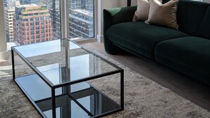Un salon moderne avec une table basse en verre, un canapé vert et une vue sur la ville grâce à de grandes fenêtres.
