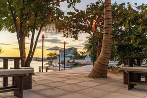 An outdoor seating area with tables and chairs, palm trees, and a view of a modern building across a body of water. - Luxury Sky Villa | Outdoor Pool | Oceanfront Family Getaway (Long Bay Hills)