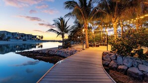 A wooden boardwalk leading to a beach area with palm trees and string lights, overlooking a calm body of water and a row of buildings. - Oceanfront Villa | Balcony | Designer 3BR with Endless Views (Long Bay Hills)