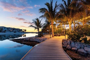A wooden pathway leading to a seating area with string lights, palm trees, and a calm body of water. - Oceanfront Villa | Balcony | Designer 3BR with Endless Views (Long Bay Hills)