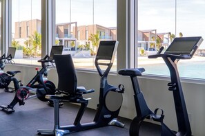 Une rangée de vélos d’appartement équipés d’écrans est disposée dans une pièce dotée de grandes fenêtres donnant sur une plage et des bâtiments modernes.