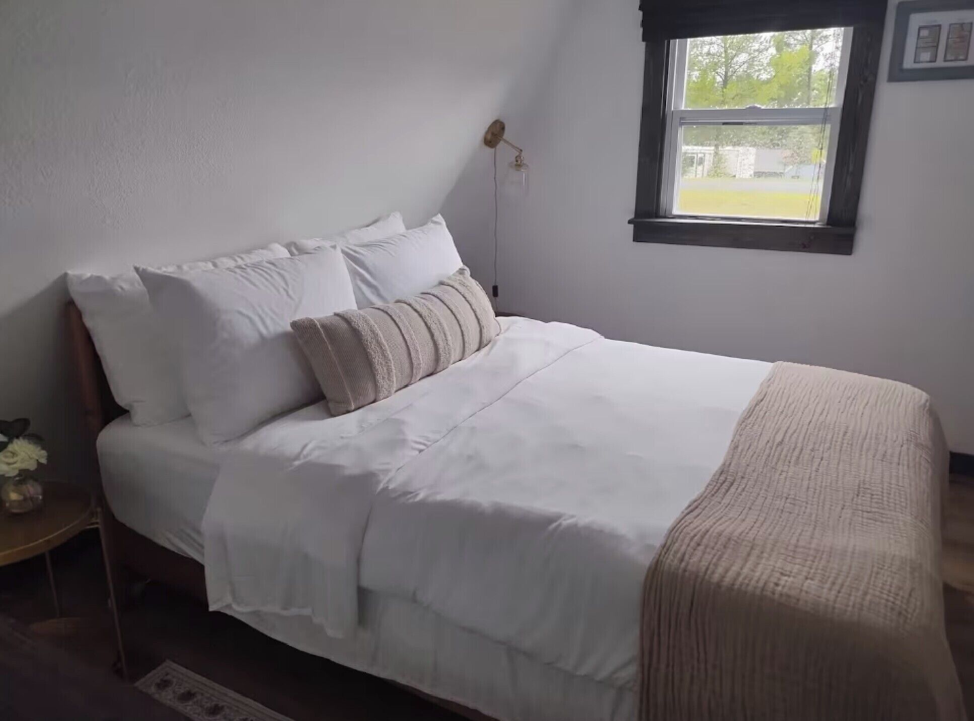 Ein ordentlich bezogenes Bett mit weißer Bettwäsche und einer beige farbenen Tagesdecke, ein Nachttisch mit einer Blumenvase und ein Fenster mit Blick auf Grün.