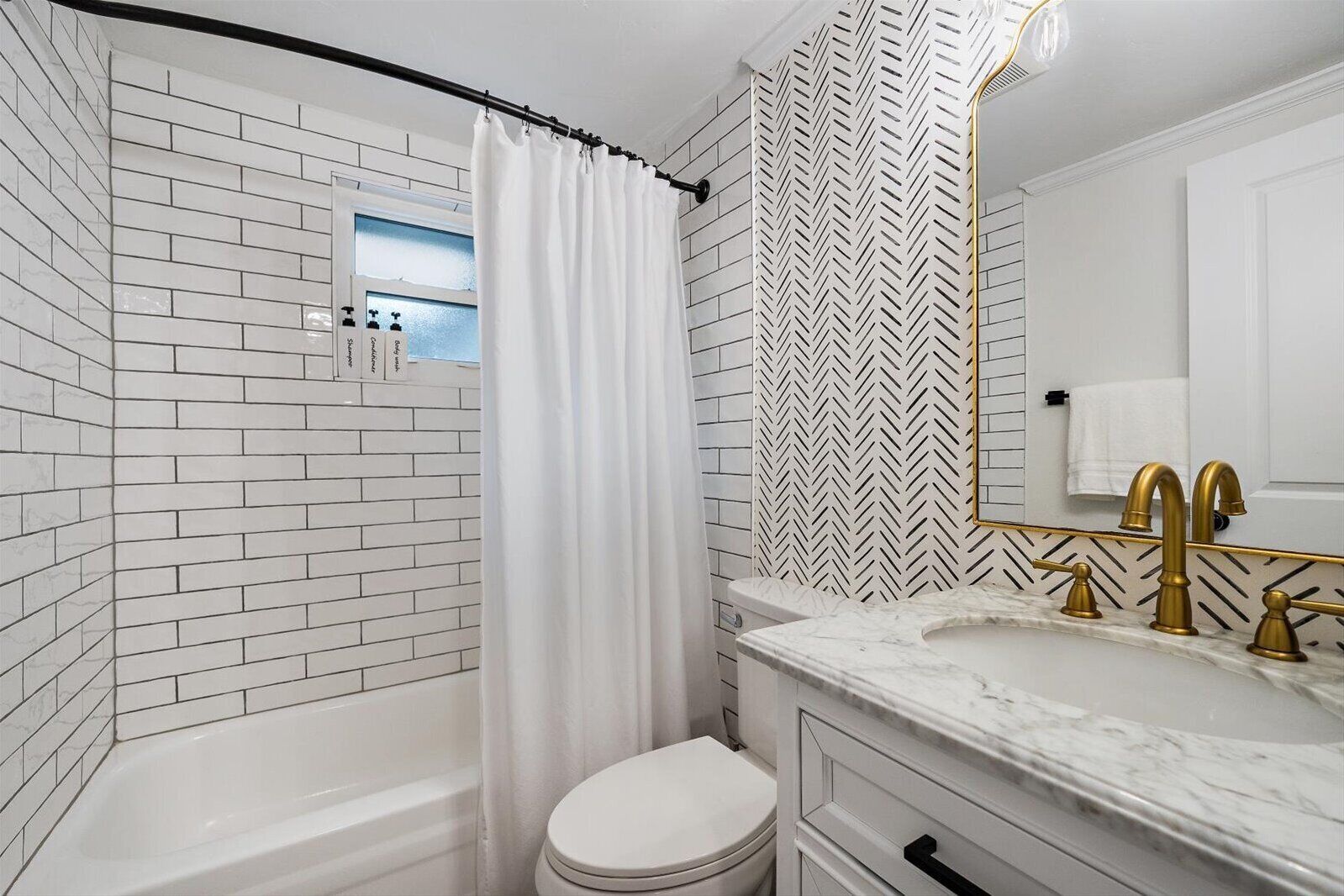 A bathroom with white brick walls, a white toilet, a shower with a white curtain, a gold-framed mirror, and a marble sink with gold faucets.