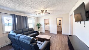 A living room with a sofa, a coffee table, a ceiling fan, a TV, and a kitchen area in the background.