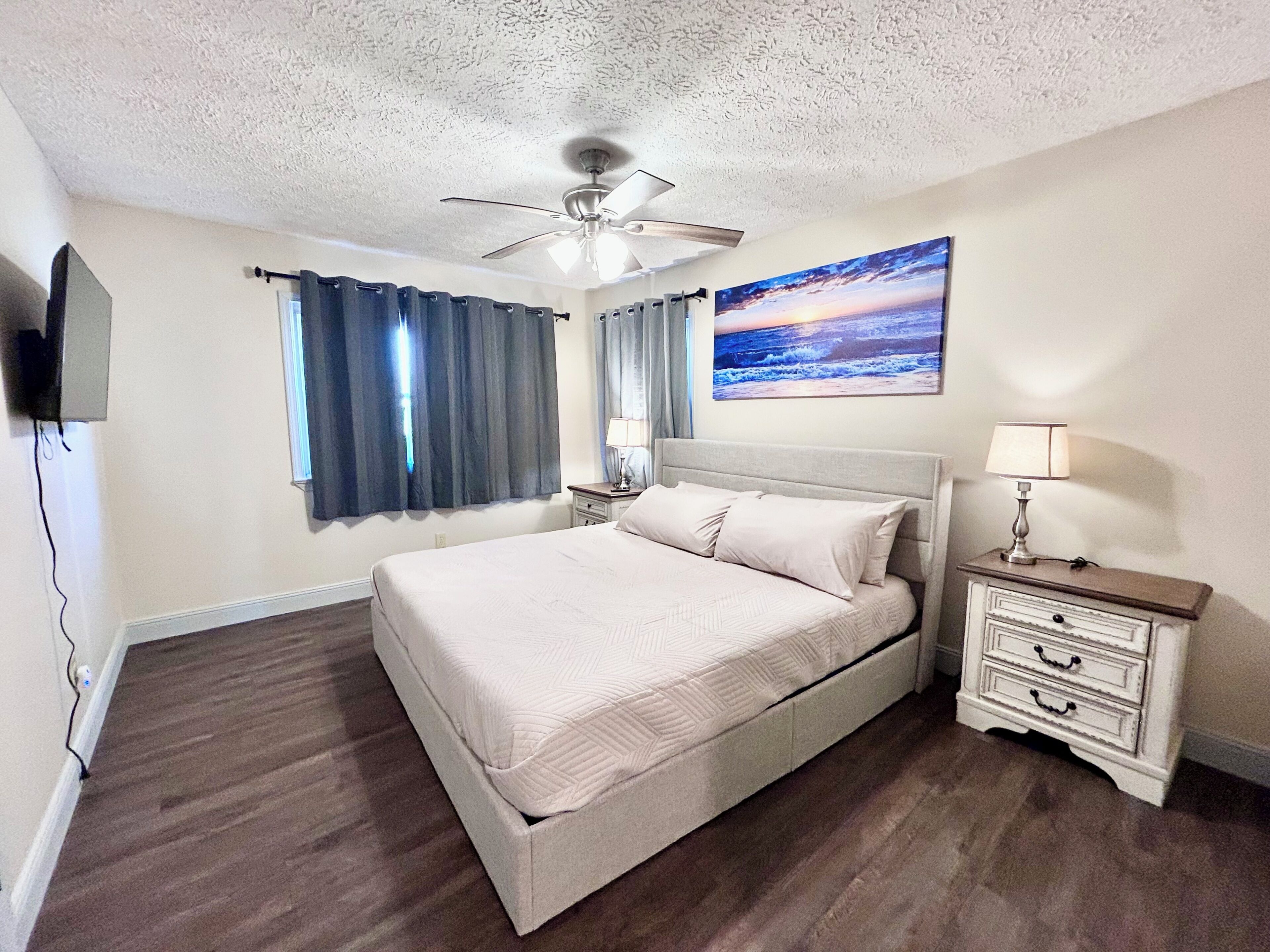 A bedroom with a bed, a nightstand, a lamp, a ceiling fan, a painting, and a TV mounted on the wall.